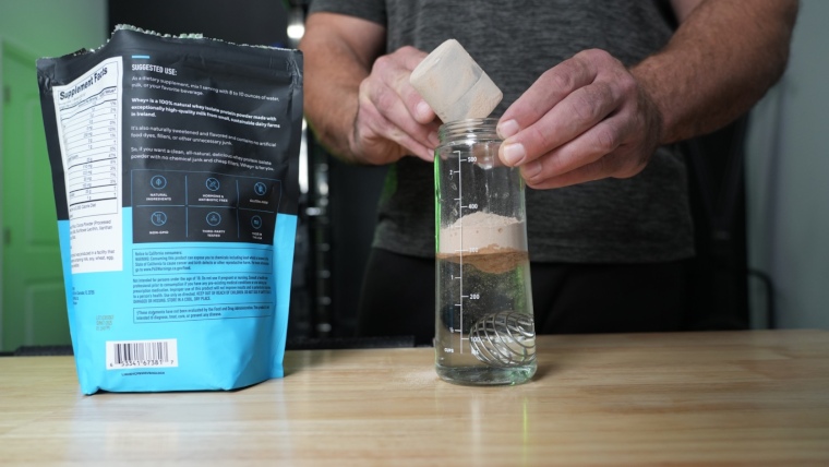 A person pours a scoop of Legion Whey protein powder into a shaker bottle of water.